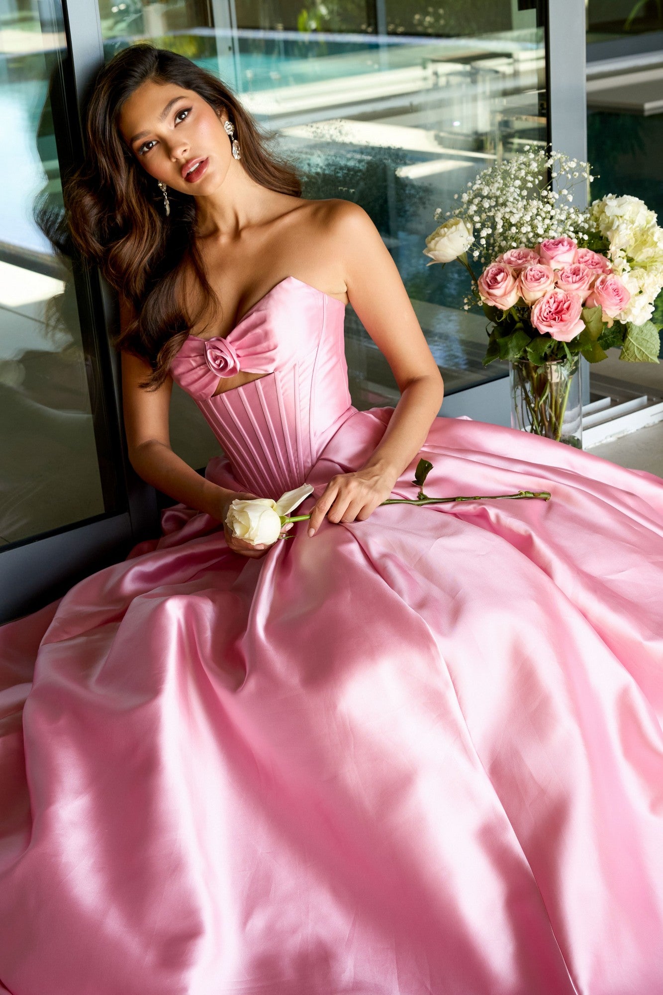 A woman in the Ava Presley 47223 Strapless Satin Prom Ball Gown sits indoors holding a white rose, with a bouquet of pink and white flowers beside her.